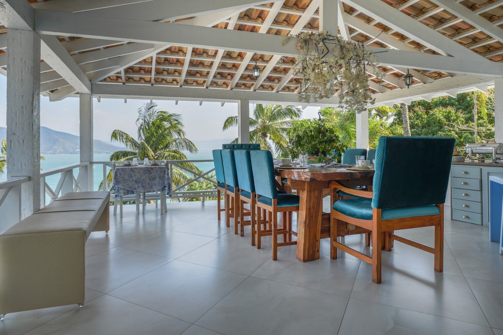 Área de jantar ao ar livre com mesa de madeira, cadeiras verde-azuladas e vista para a montanha através de colunas brancas brilhantes.