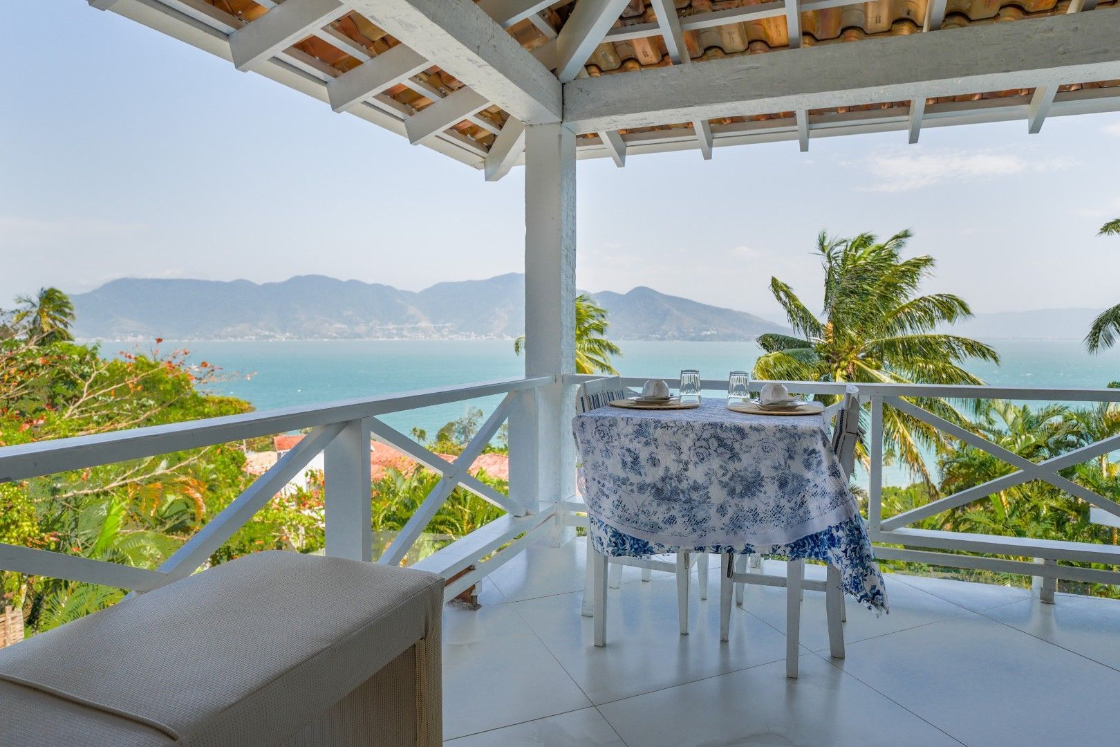 Varanda com mesa e cadeiras com vista para a água turquesa, montanhas e palmeiras.