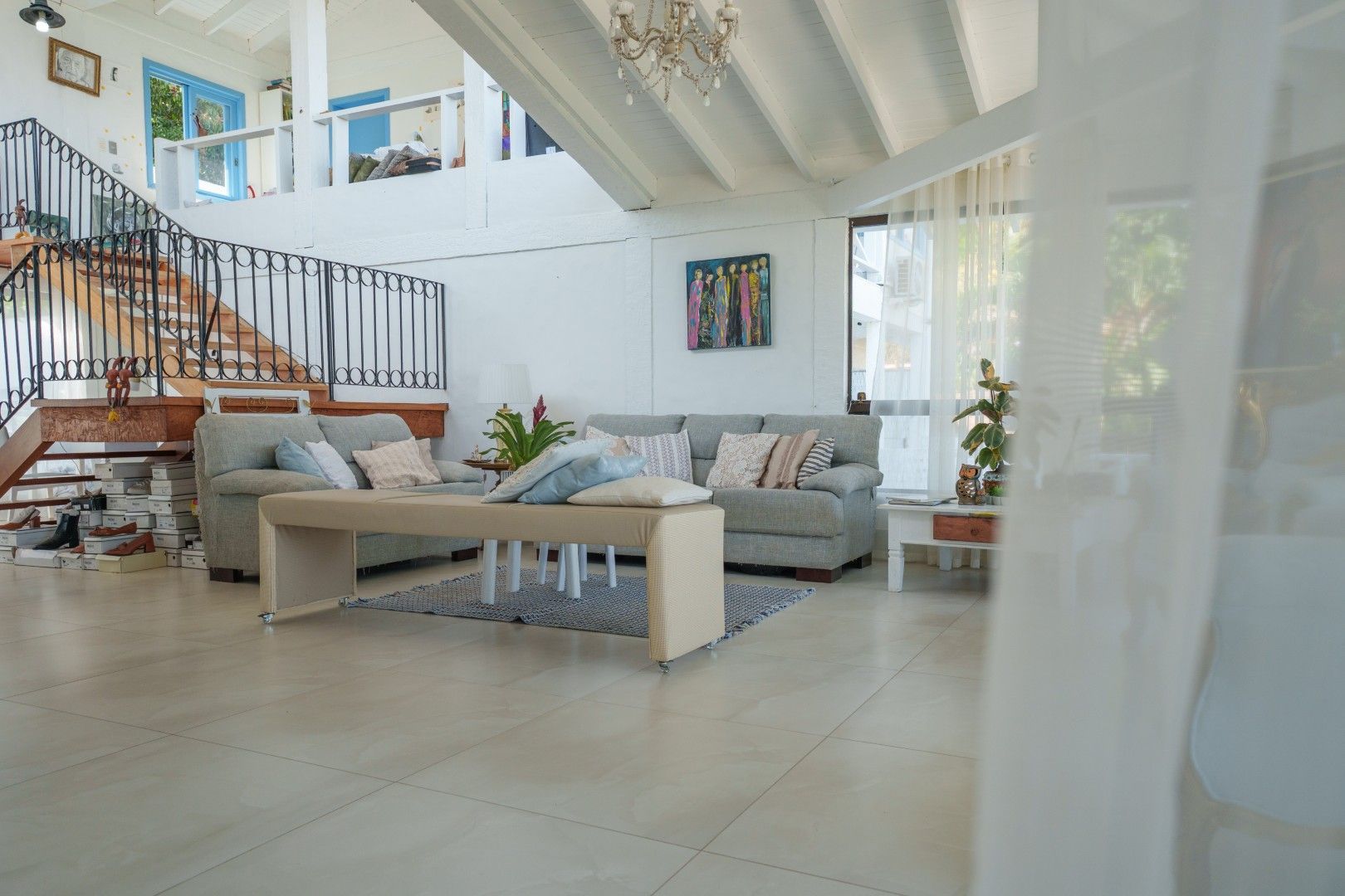 Sala de estar moderna e iluminada com sofá cinza, mesa de centro creme, escada e janelas amplas.