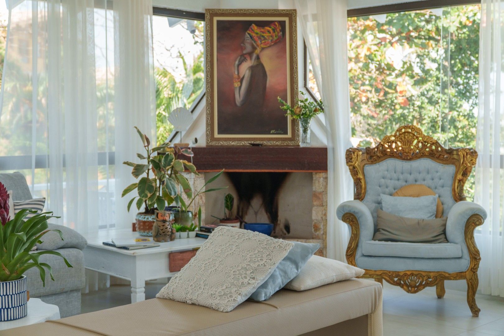Sala de estar elegante com chaise bege, cadeira dourada ornamentada, lareira e janelas ensolaradas com plantas.