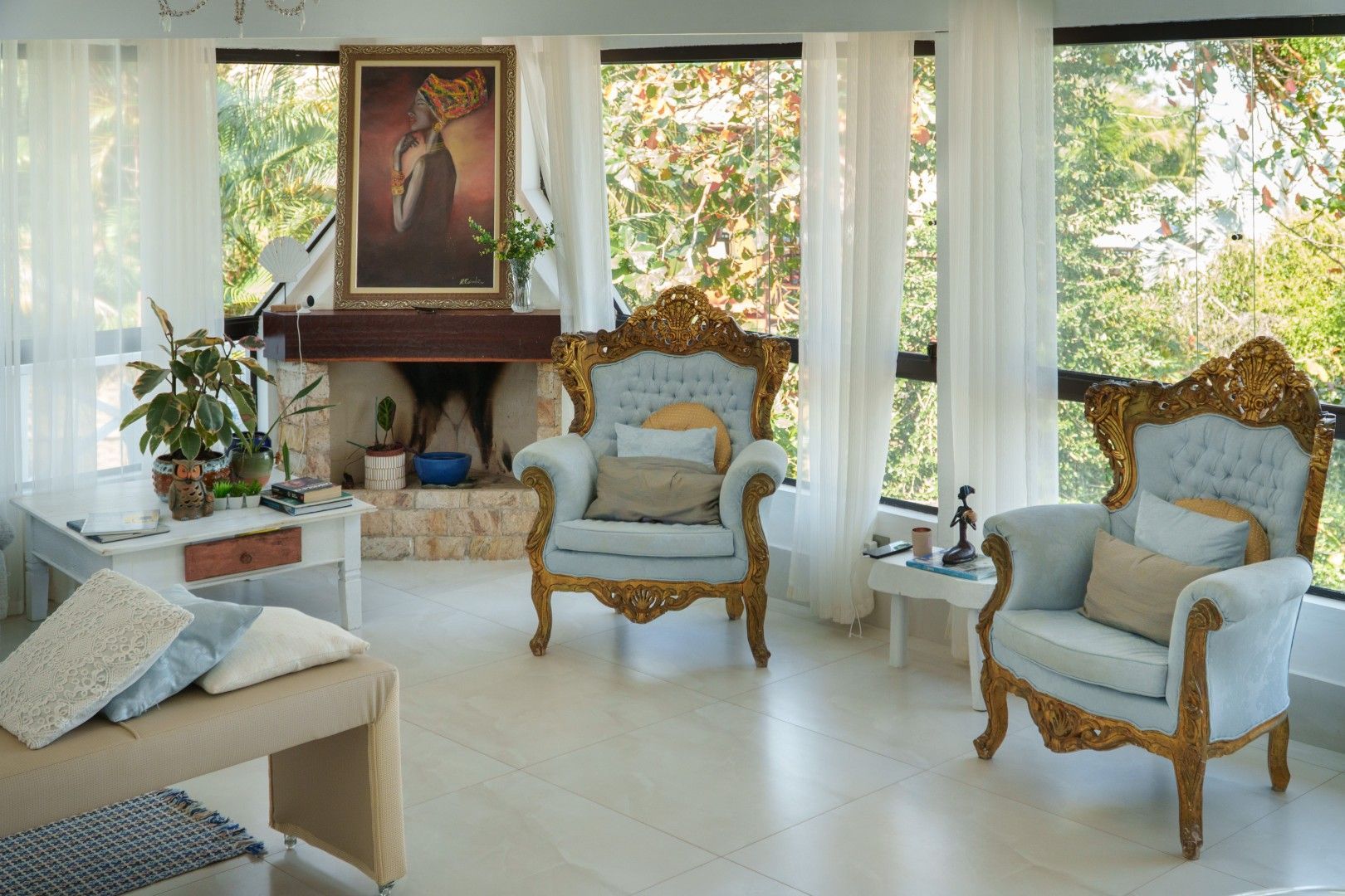 Sala de estar elegante e branca com cadeiras ornamentadas em dourado, lareira e grandes janelas com vista para a vegetação.