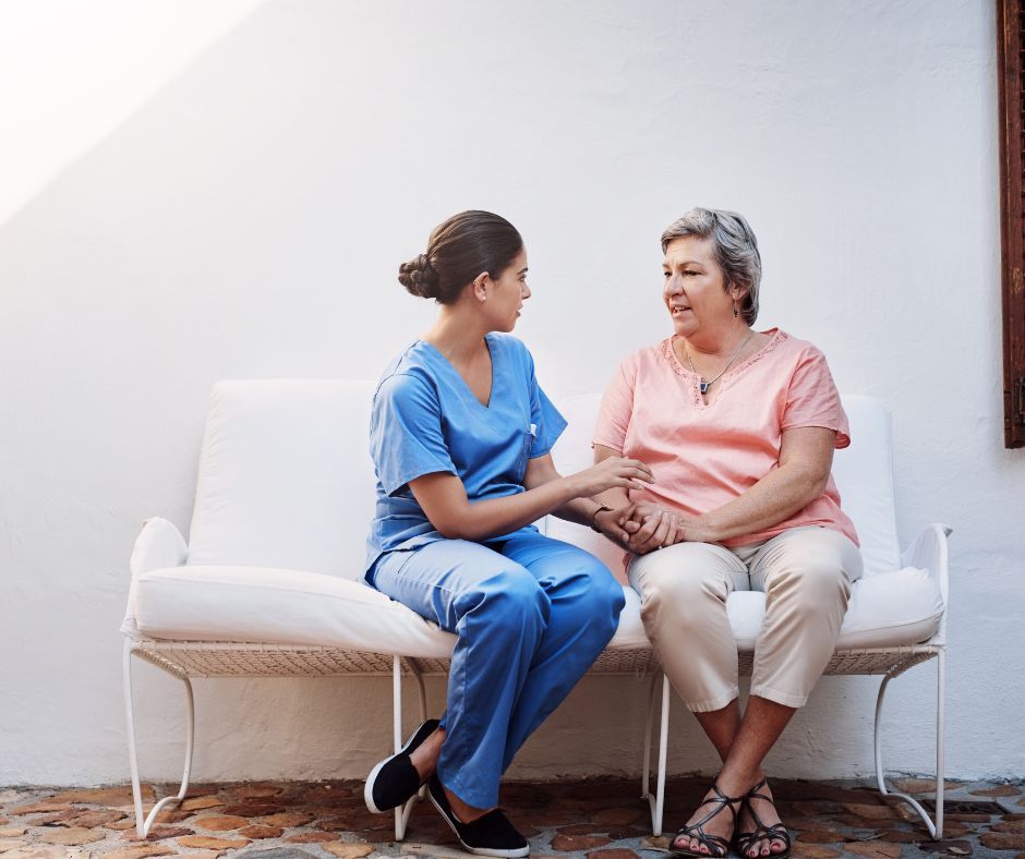 Nurse in blue scrubs speaking with an older woman on a bench.