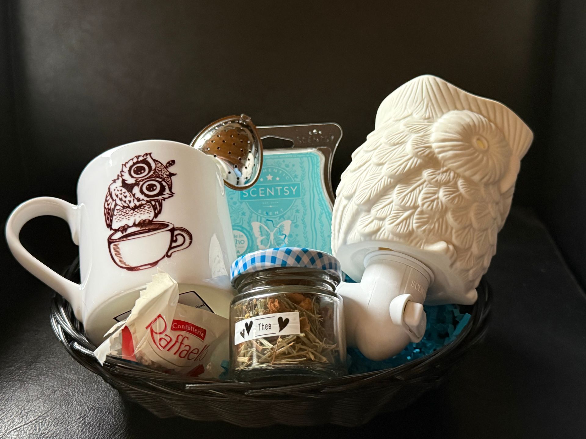 Een geschenkmand met een uilenmok, thee, kaars, uilenwarmer en theezeefje.