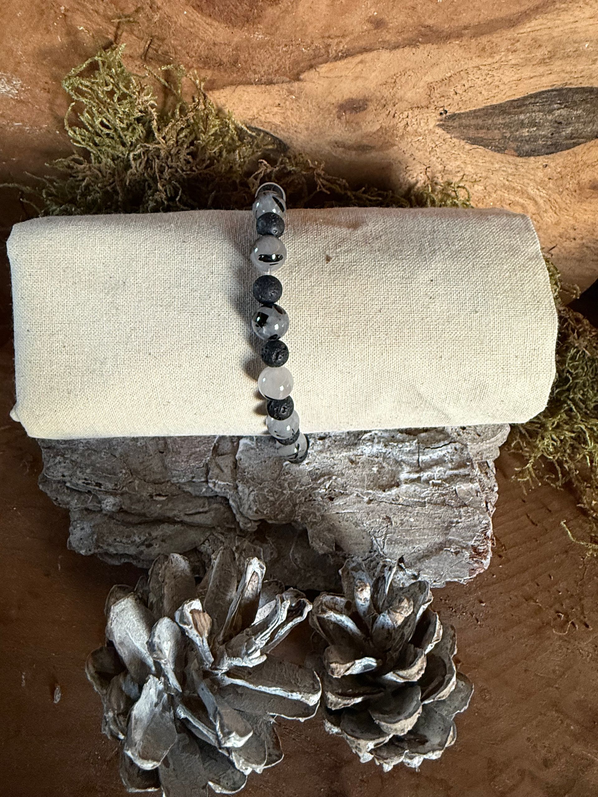 Armband met zwart-witte kralen op een opgerolde, crèmekleurige stof, bevestigd op hout en versierd met dennenappels.