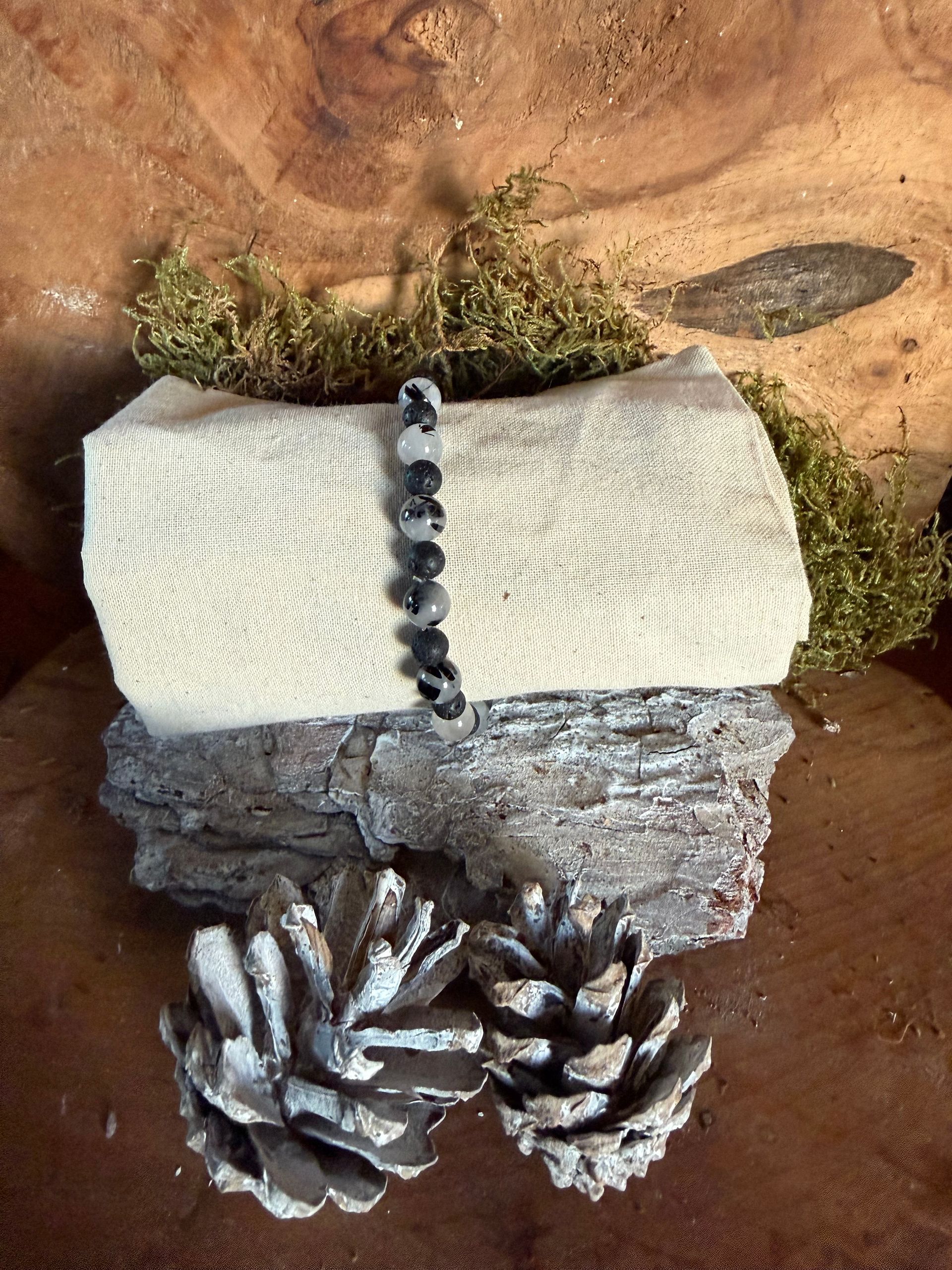 Armband met zwart-witte kralen op een opgerolde, crèmekleurige stof, bevestigd op hout en versierd met dennenappels.