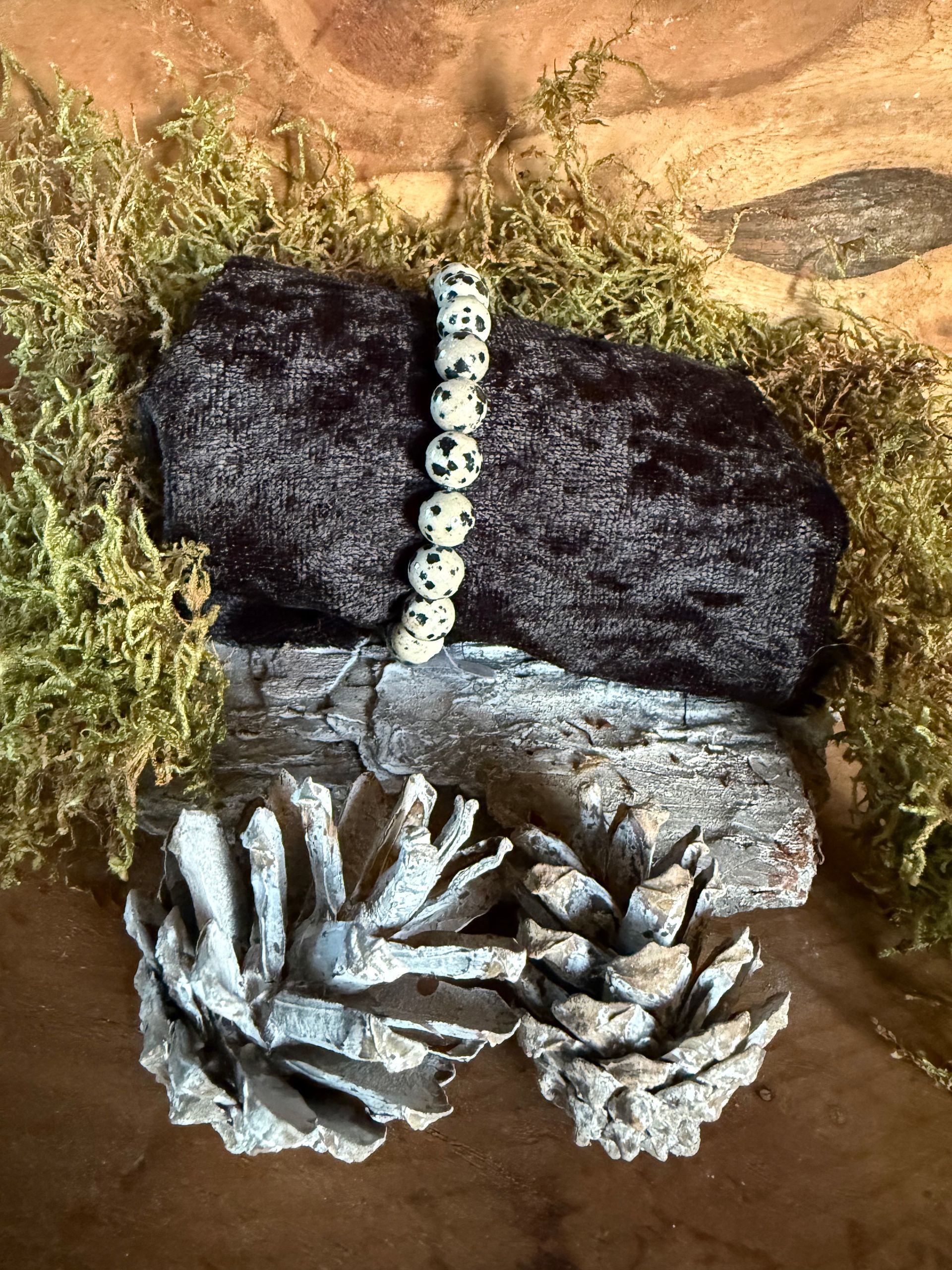 Een kralenarmband op een donkere, gespikkelde steen, bovenop een lichtgekleurde steen met mos en twee dennenappels.