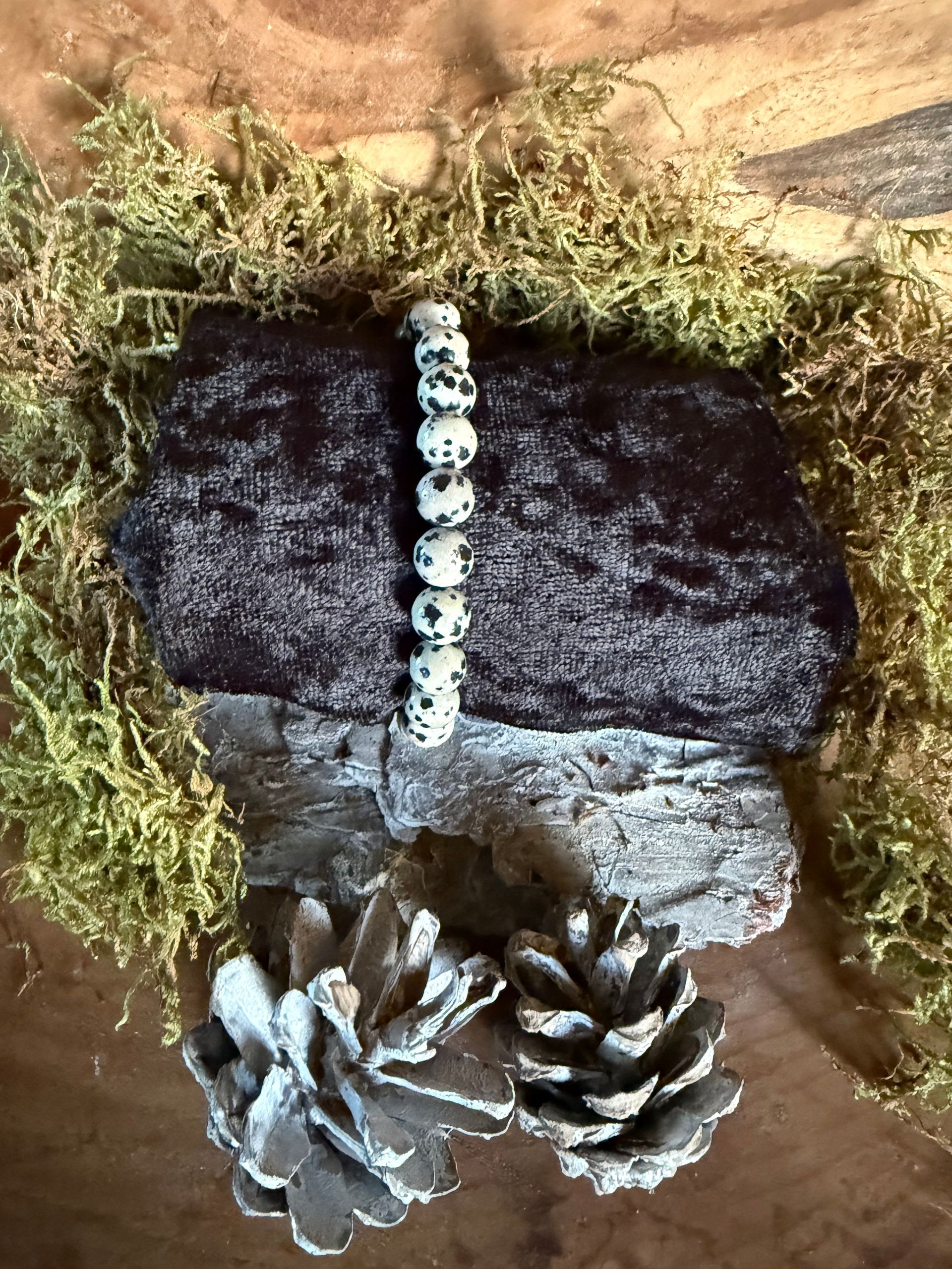 Een kralenarmband op een donkere, gespikkelde steen, bovenop een lichtgekleurde steen met mos en twee dennenappels.