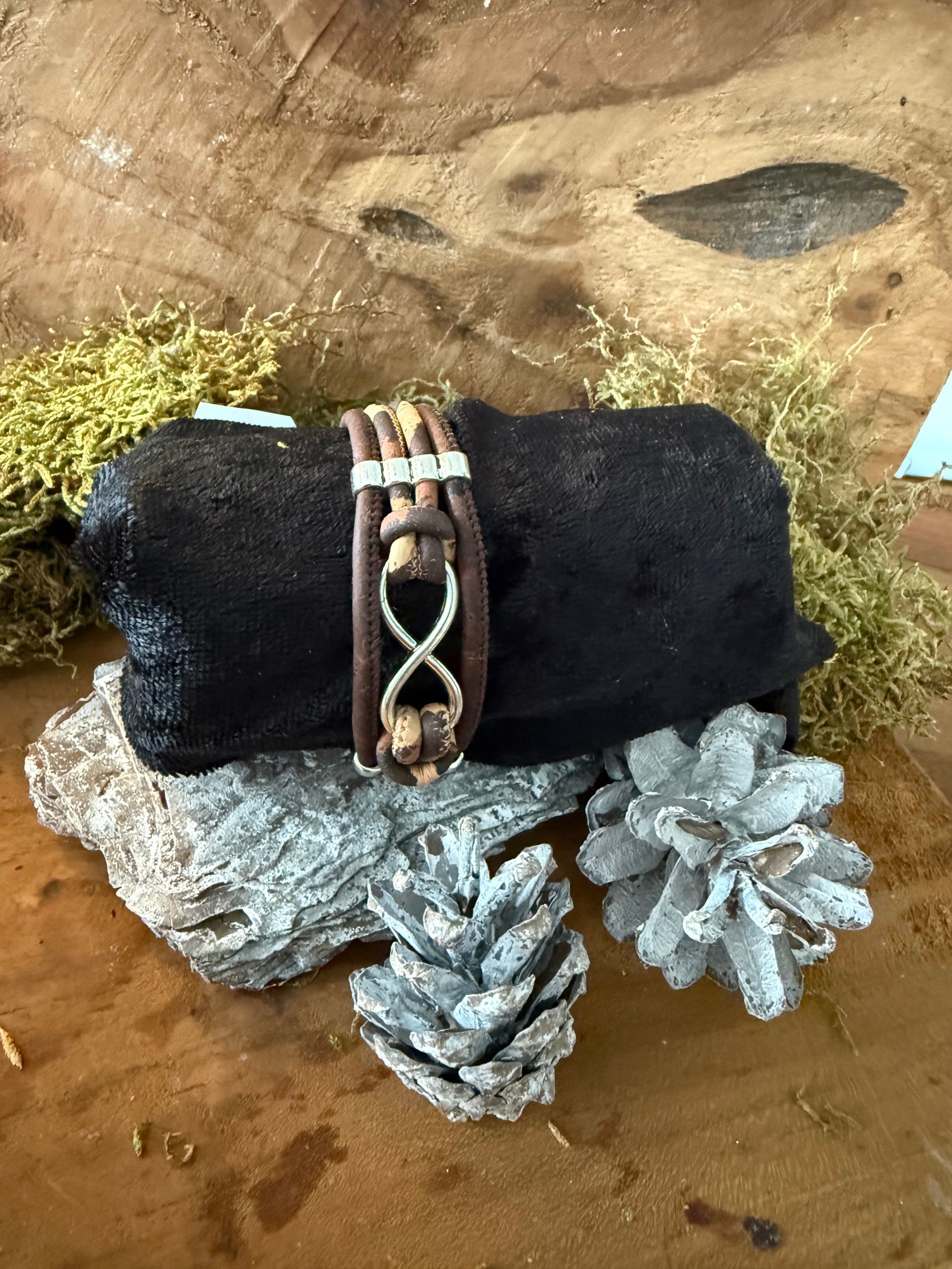 Leren armband met een oneindigheidssymbool, afgebeeld op een donkere boomstam, omringd door dennenappels en mos.