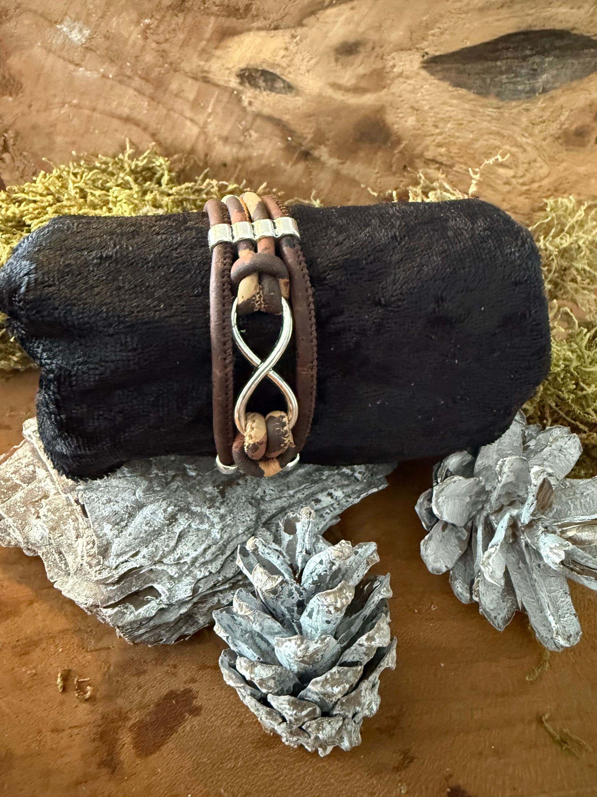 Bruine leren armband met een zilveren oneindigheidssymbool op een zwarte boomstam, versierd met dennenappels.