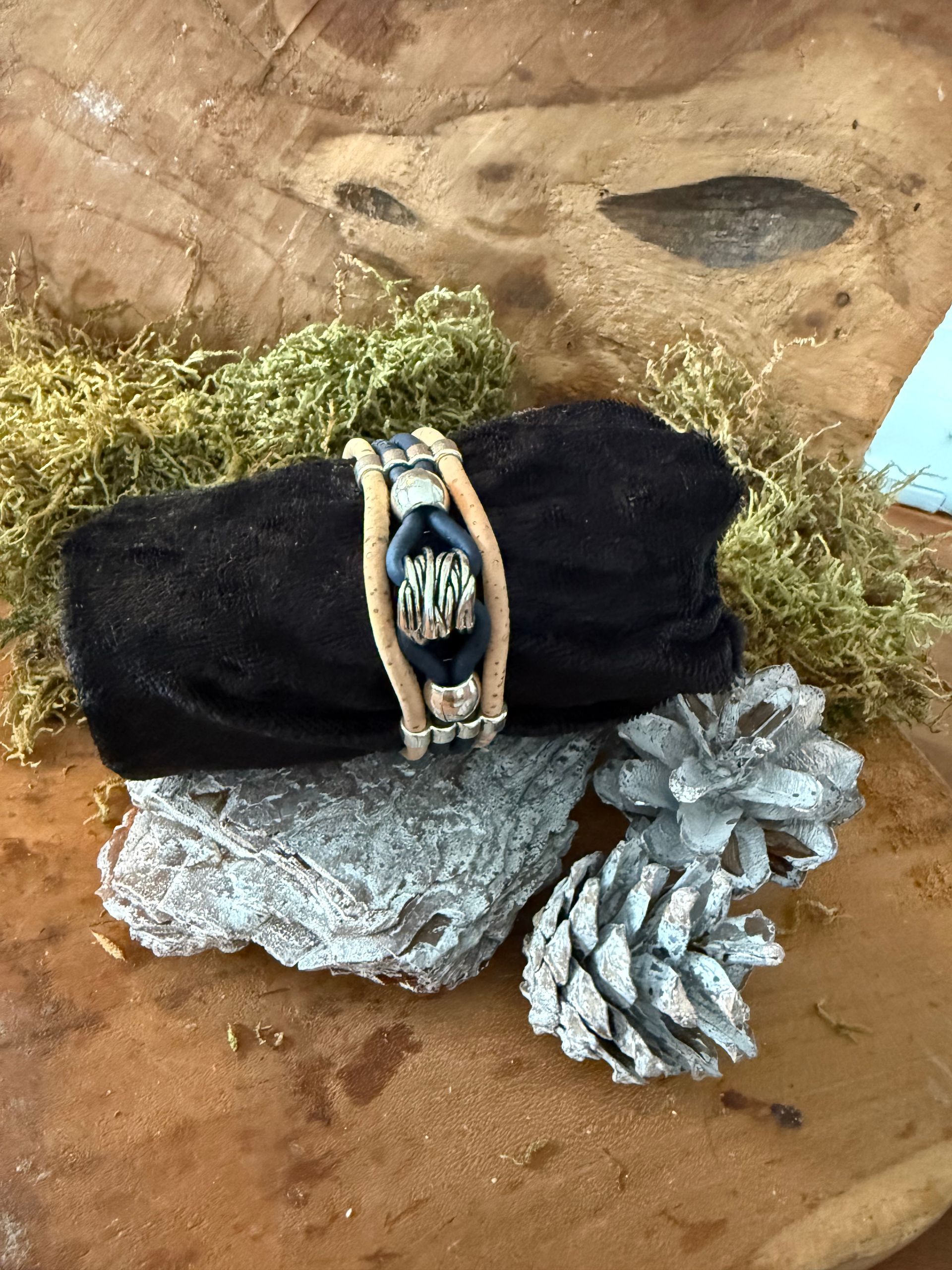 Armband op een zwart doek op een houten schijf met mos, beschilderde steentjes en dennenappels.