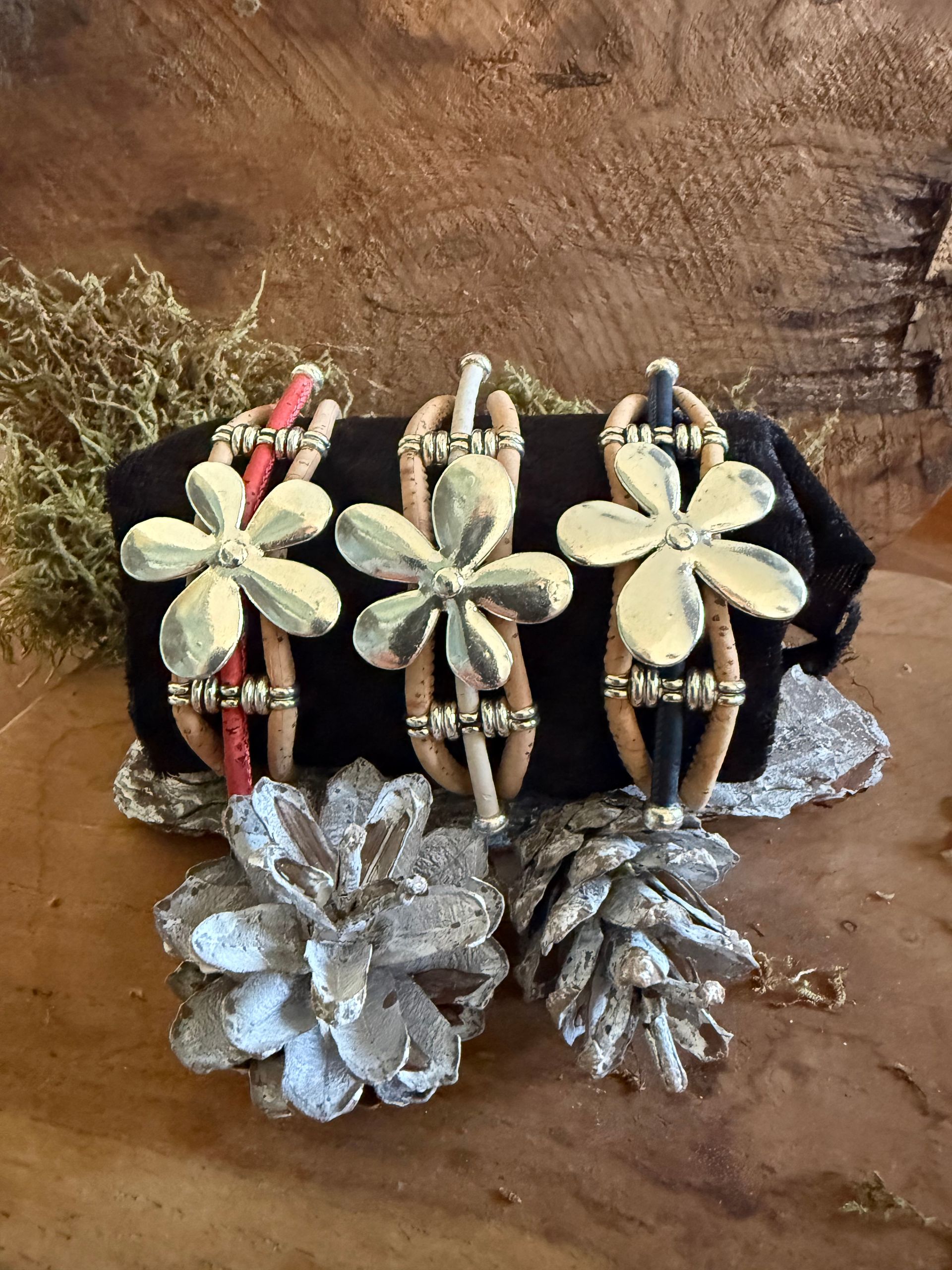 Drie bloemvormige armbanden met accenten liggen op een zwart kleed met zilveren dennenappels, op een houten ondergrond.