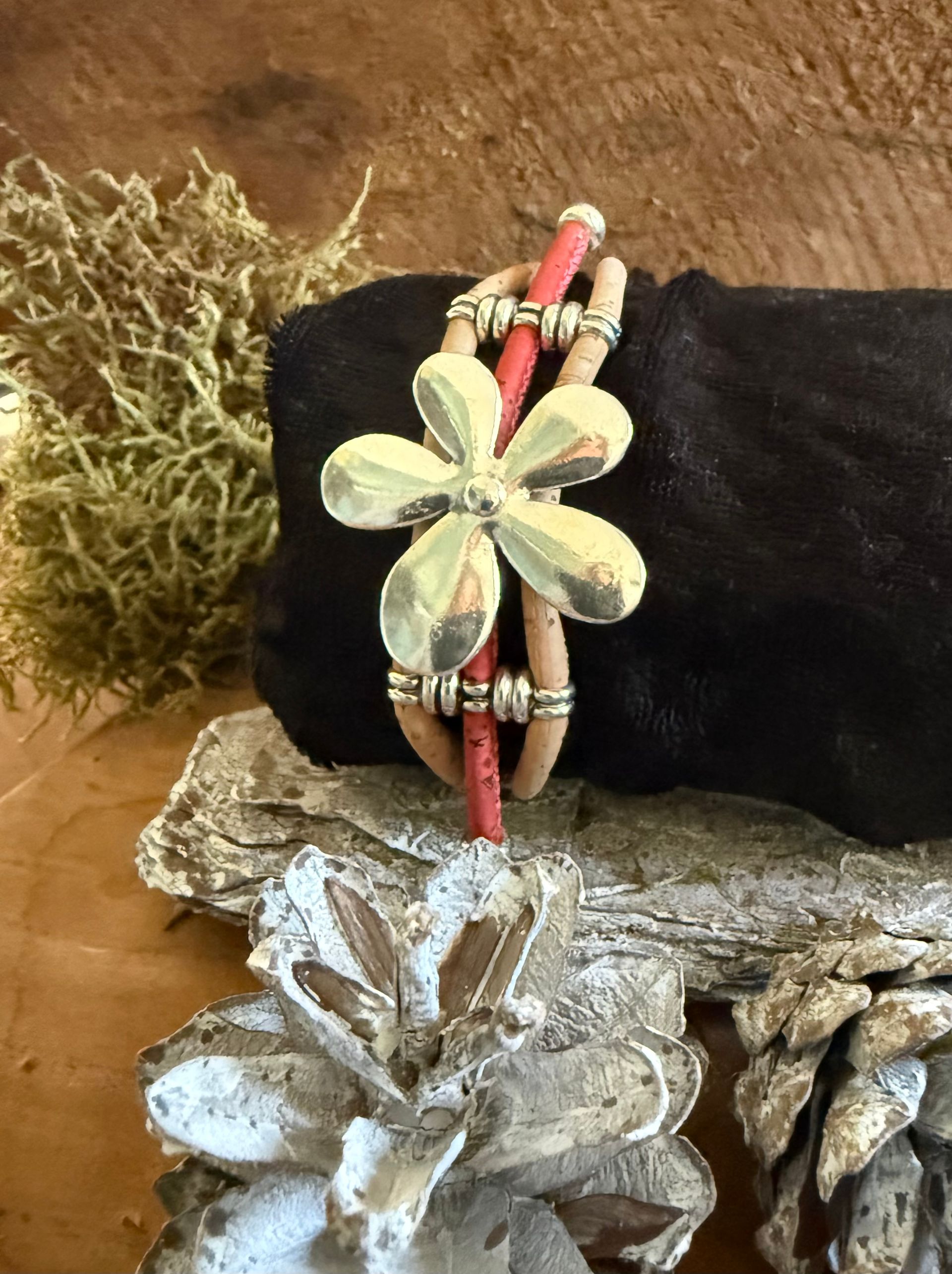 Armband met zilveren bloemhanger, rood koord en kurkdetails, gepresenteerd op een rustieke ondergrond met dennenappels.