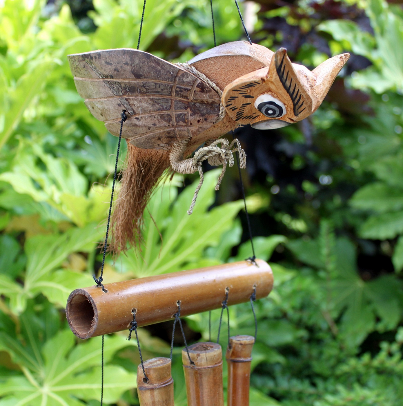 Een windgong in de vorm van een uil in een tuin. Bruine en beige kleuren. Bamboe buizen hangen eronder.
