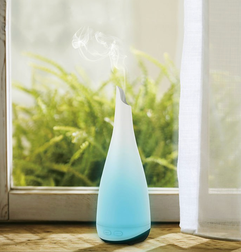 Witte aromadiffuser die nevel verspreidt, op een houten oppervlak met twee bovenliggende lampen.