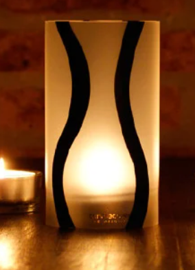 Een brandende kaars in een witte, rechthoekige houder met zwarte, gebogen accenten.