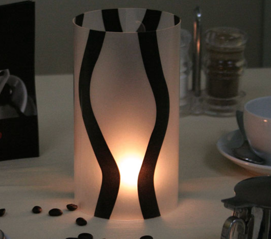 Een brandende kaars in een witte, rechthoekige houder met zwarte, gebogen accenten.