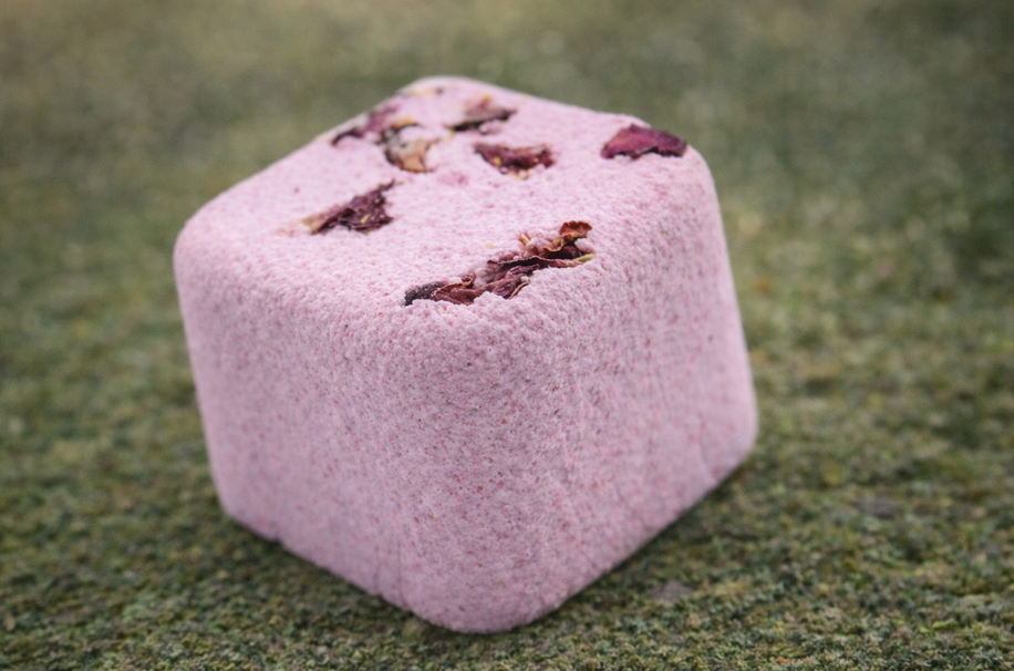 Roze, kubusvormige bruisbal voor in bad met gedroogde rozenblaadjes op een groene, gestructureerde achtergrond.