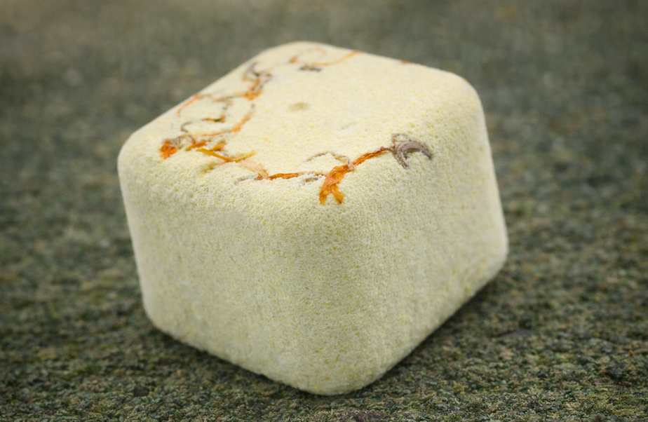 Vierkante, lichtgele stenen kubus met oranje aderen op een gestructureerd, grijsgroen oppervlak.