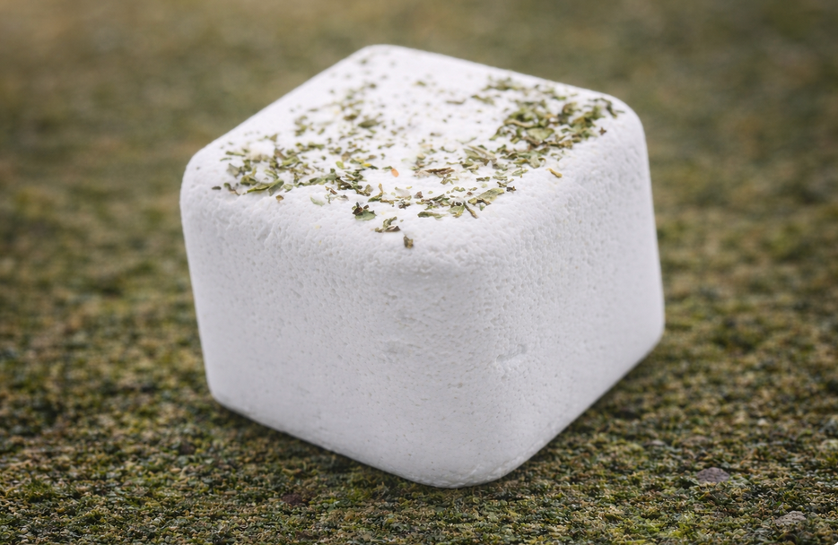 Wit vierkant dessert bestrooid met groene kruiden op een bedje van groene kruimels.