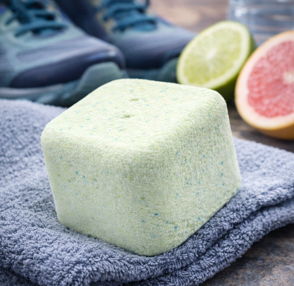 Vierkant groen schoendeodorant op een grijze handdoek, met hardloopschoenen en citrusvruchten op de achtergrond.