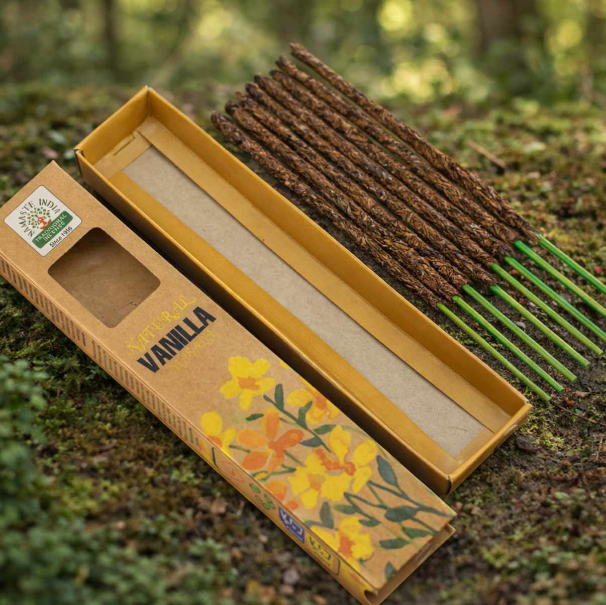 Een doosje vanillewierookstokjes met afbeeldingen van gele bloemen, geplaatst op een met mos bedekte bosbodem.