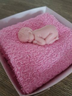 Een babyfiguurtje ligt opgerold op een roze handdoek in een witte doos.