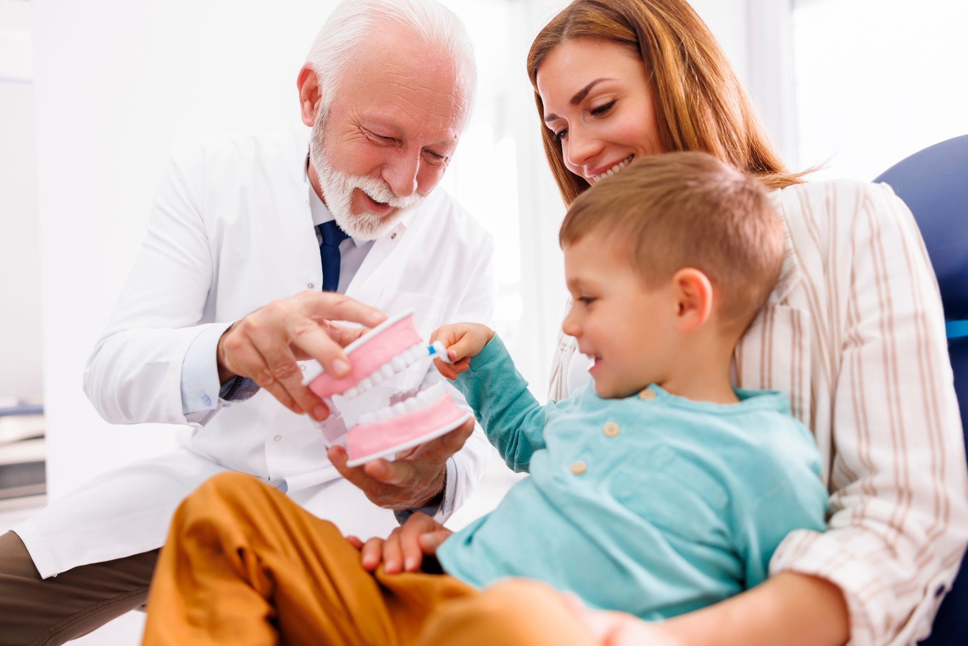 A male dentist is educating a child patient sitting in his mother's lap using a plastic tooth model. A male dentist is educating a child patient sitting in his mother's lap using a plastic tooth model.