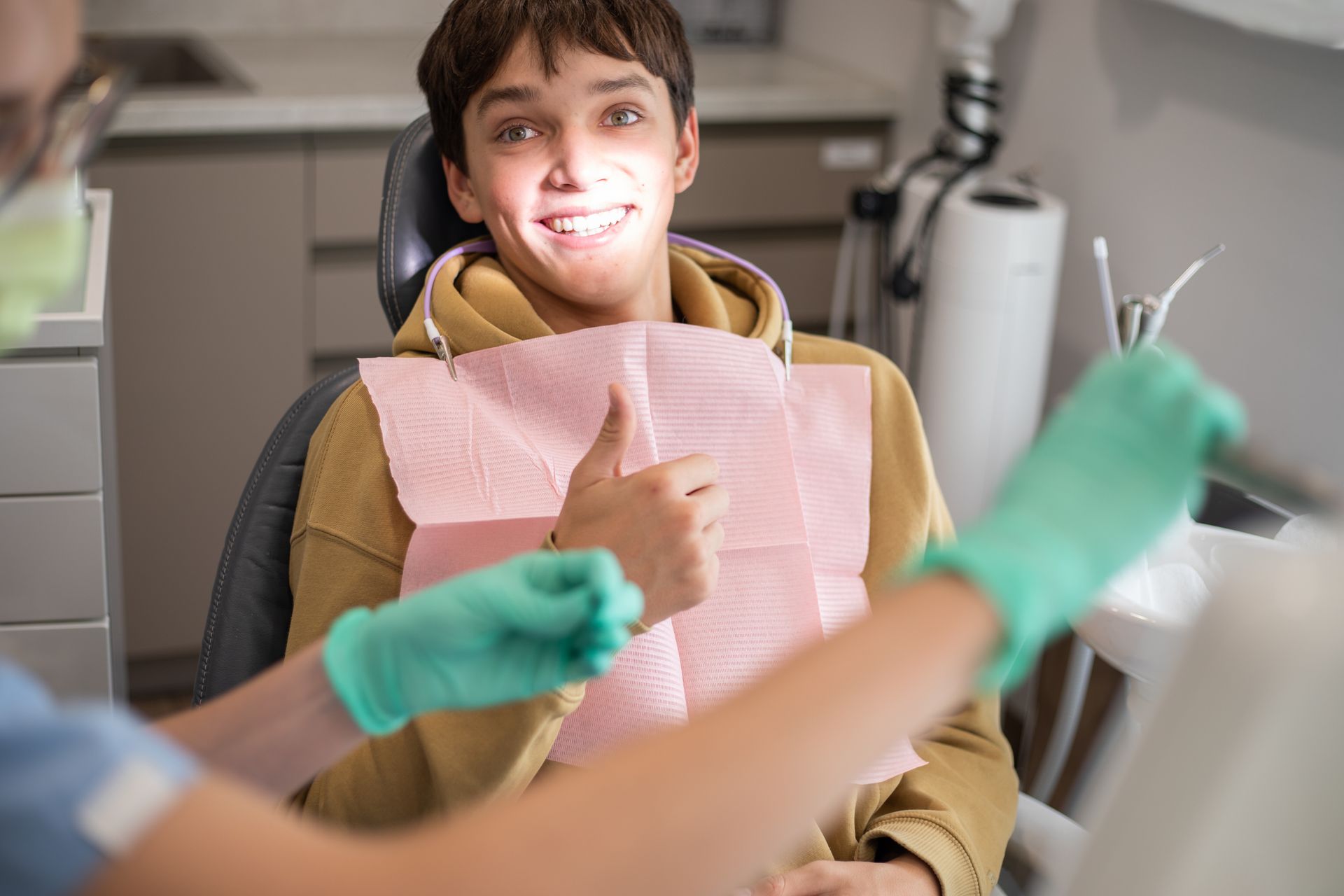 A cheerful patient in a dental chair gives a thumbs up while a dental nurse prepares tools.