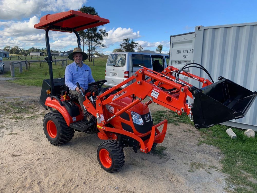 A Man is Sitting on a Small Orange Tractor With a Bucket Attached to It — Stockers & Partridge In Maitland, NSW