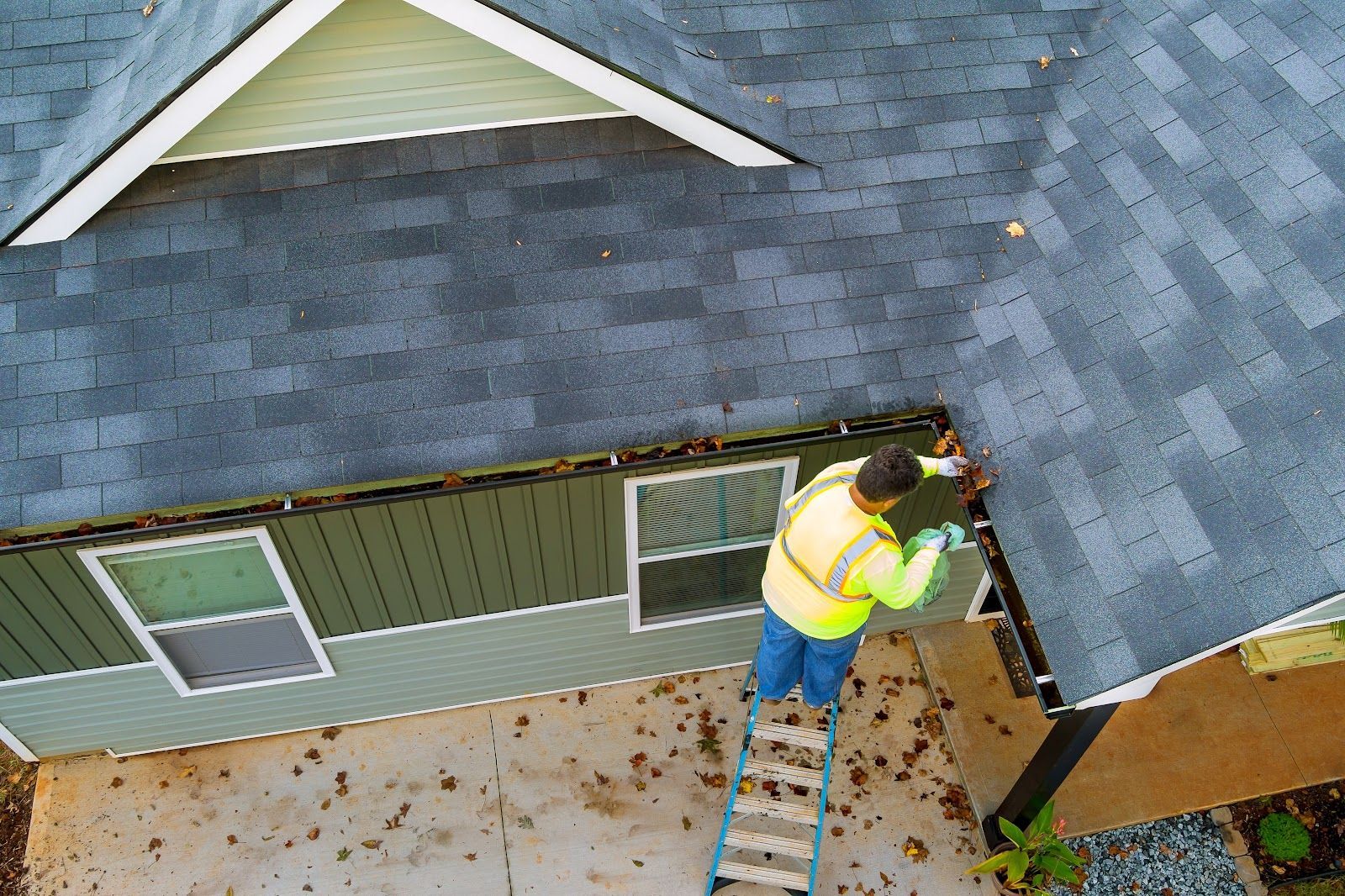 Man on ladder cleaning gutters of a house. He's wearing a safety vest. Leaves on ground.