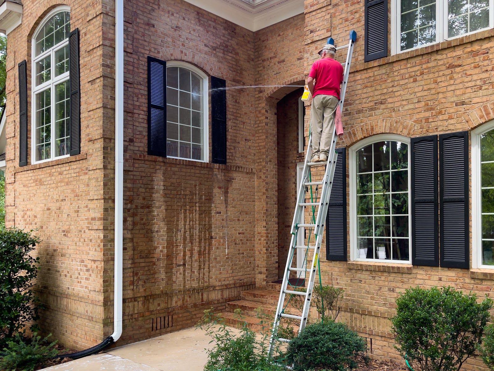 Person on ladder pressure washing brick house exterior.