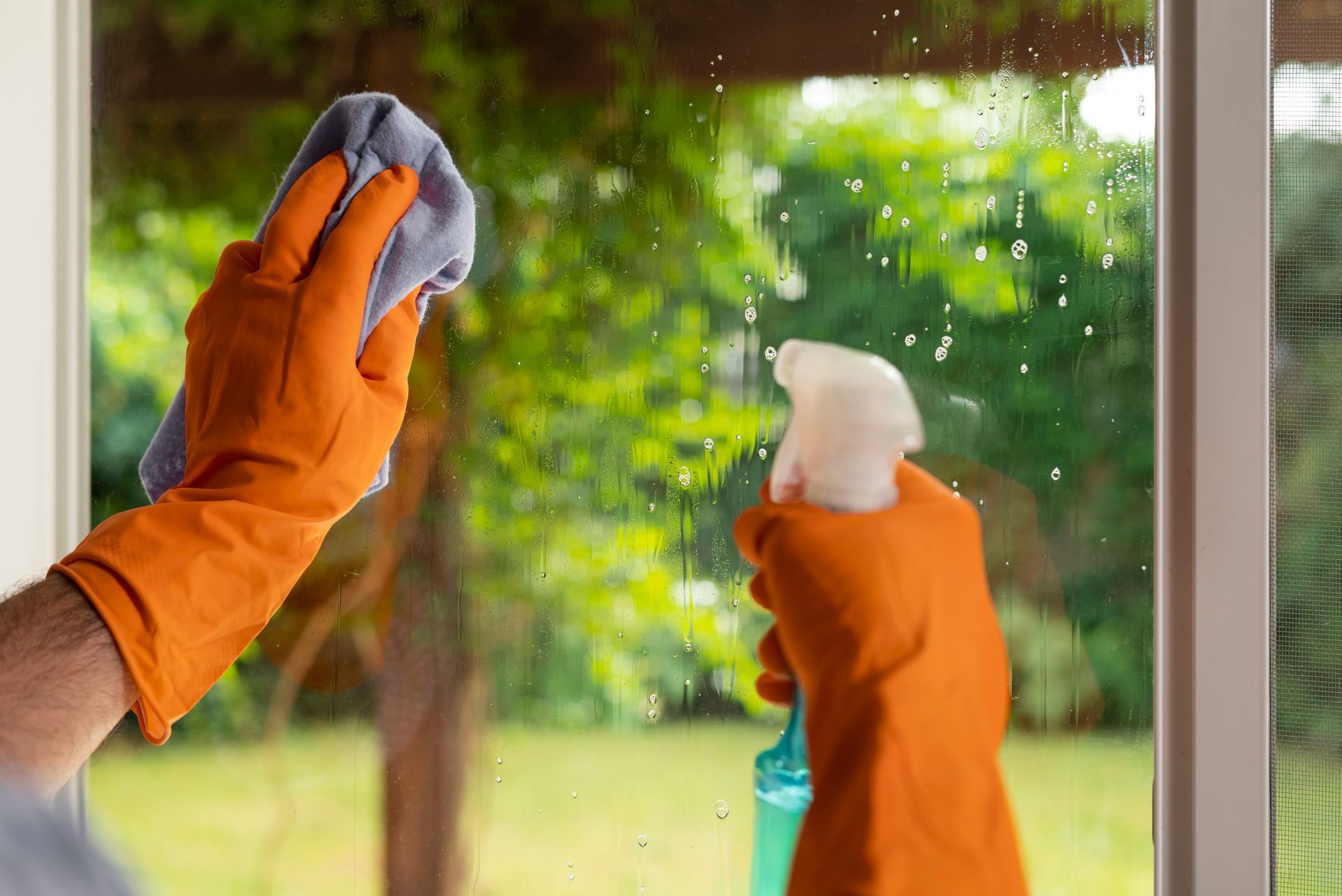 Person wearing orange gloves sprays and wipes a window clean with a cloth.