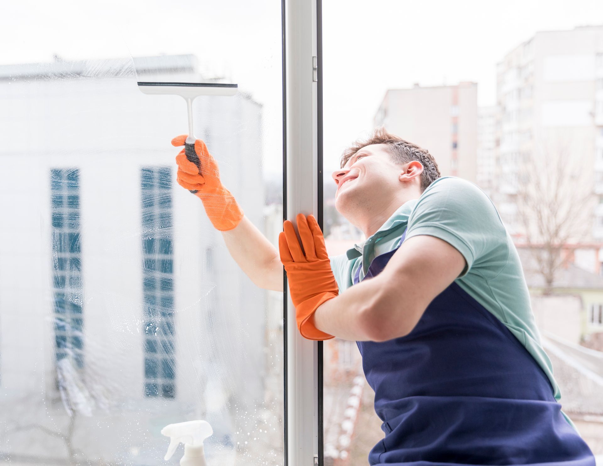 Man wearing gloves cleans a window with a squeegee, smiling, outside.