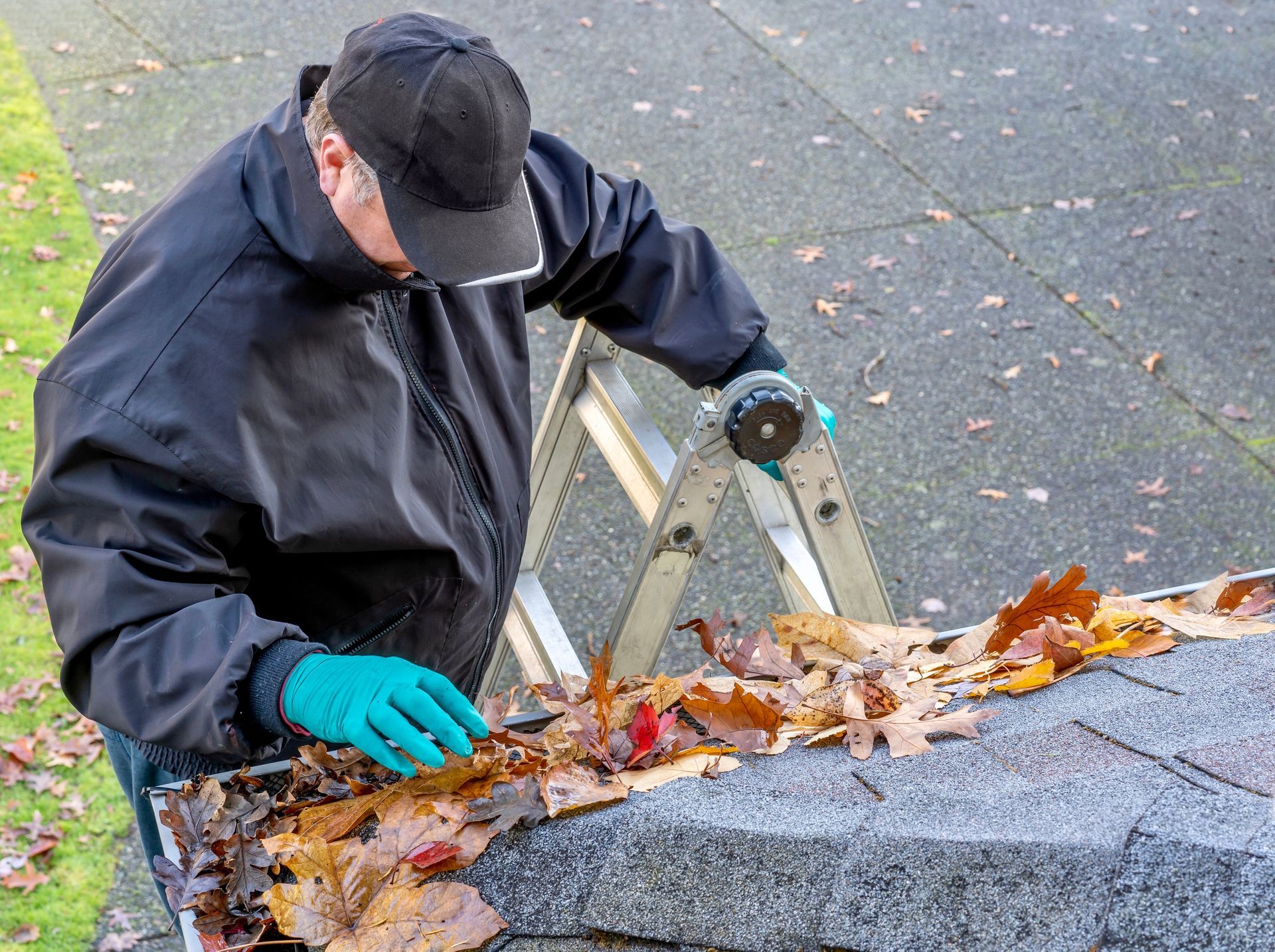 Man on ladder cleaning leaves from a gutter. Wearing a black jacket, cap, and gloves.