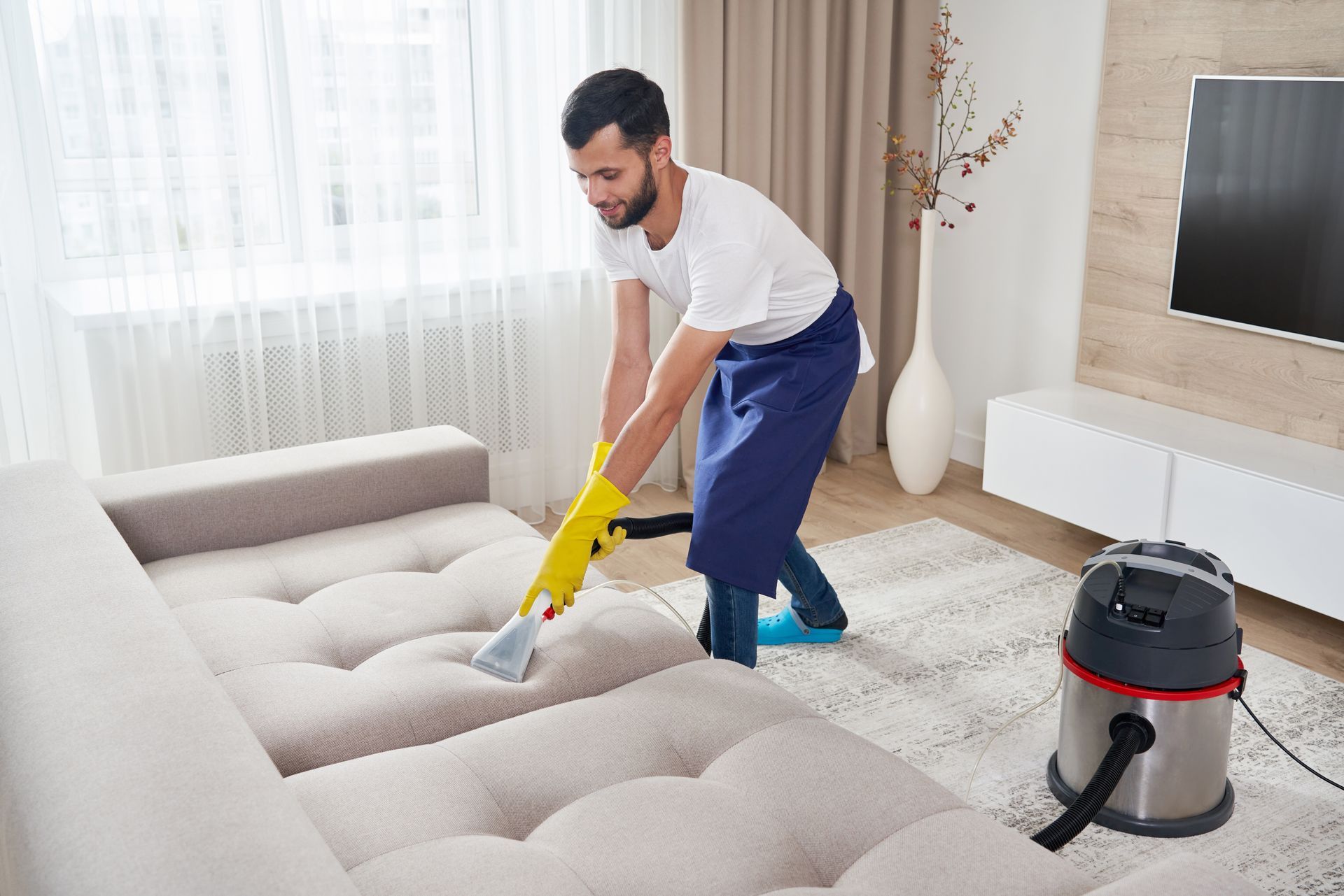 Person in yellow gloves vacuuming a light-colored sofa in a living room with a rug and television.
