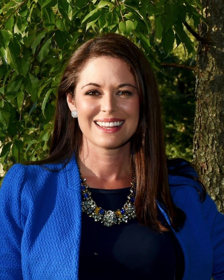 Mindy Coleman, Vice President, standing in front of green foliage.