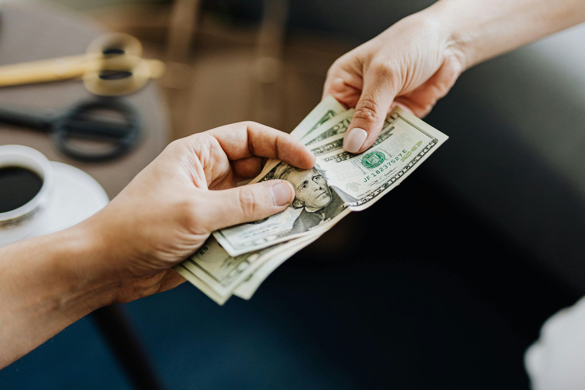 Two hands exchanging a small stack of US dollar bills in a dimly lit, indoor setting.