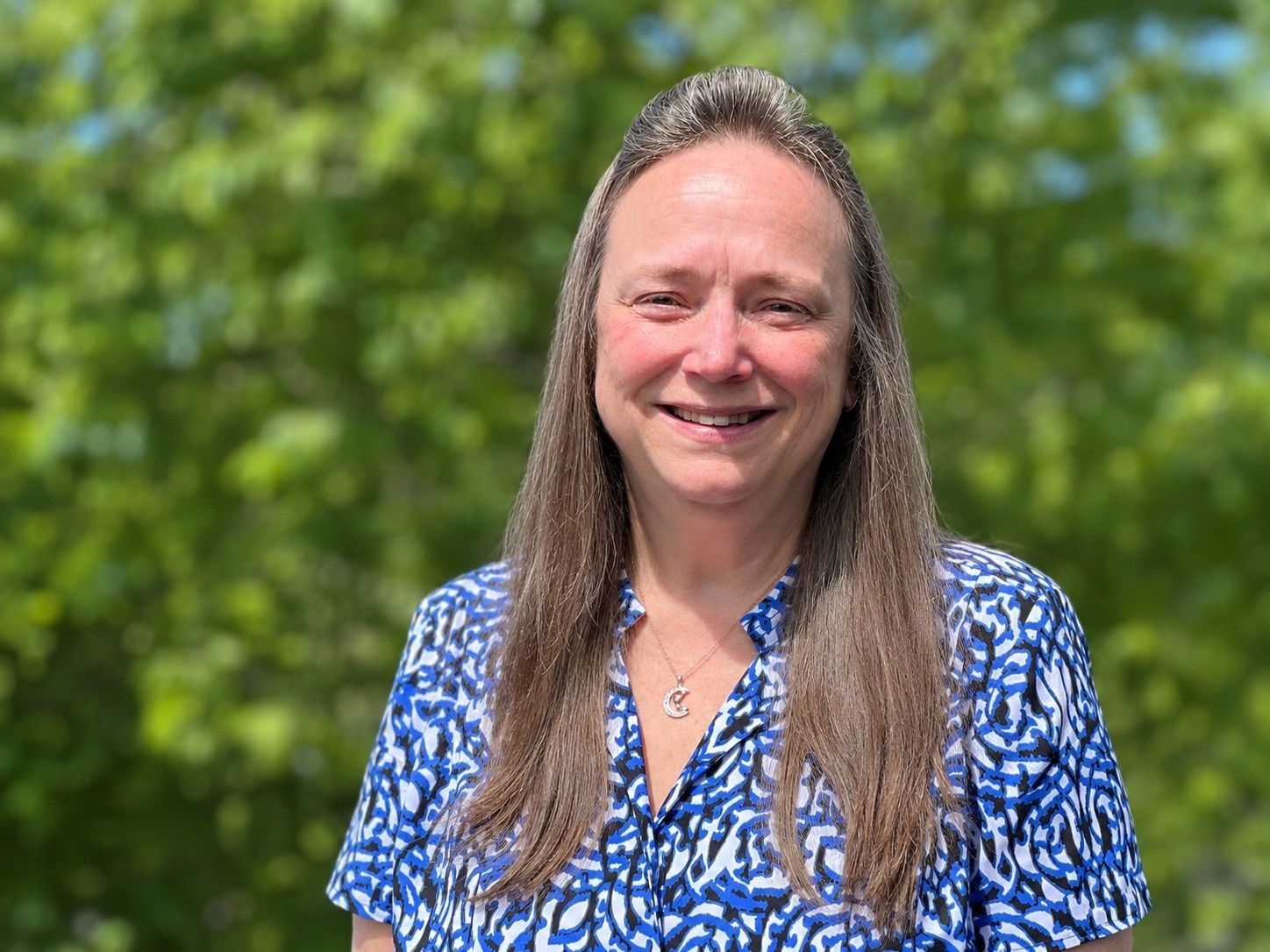 Smiling woman in a blue patterned blouse standing outdoors against a leafy green background