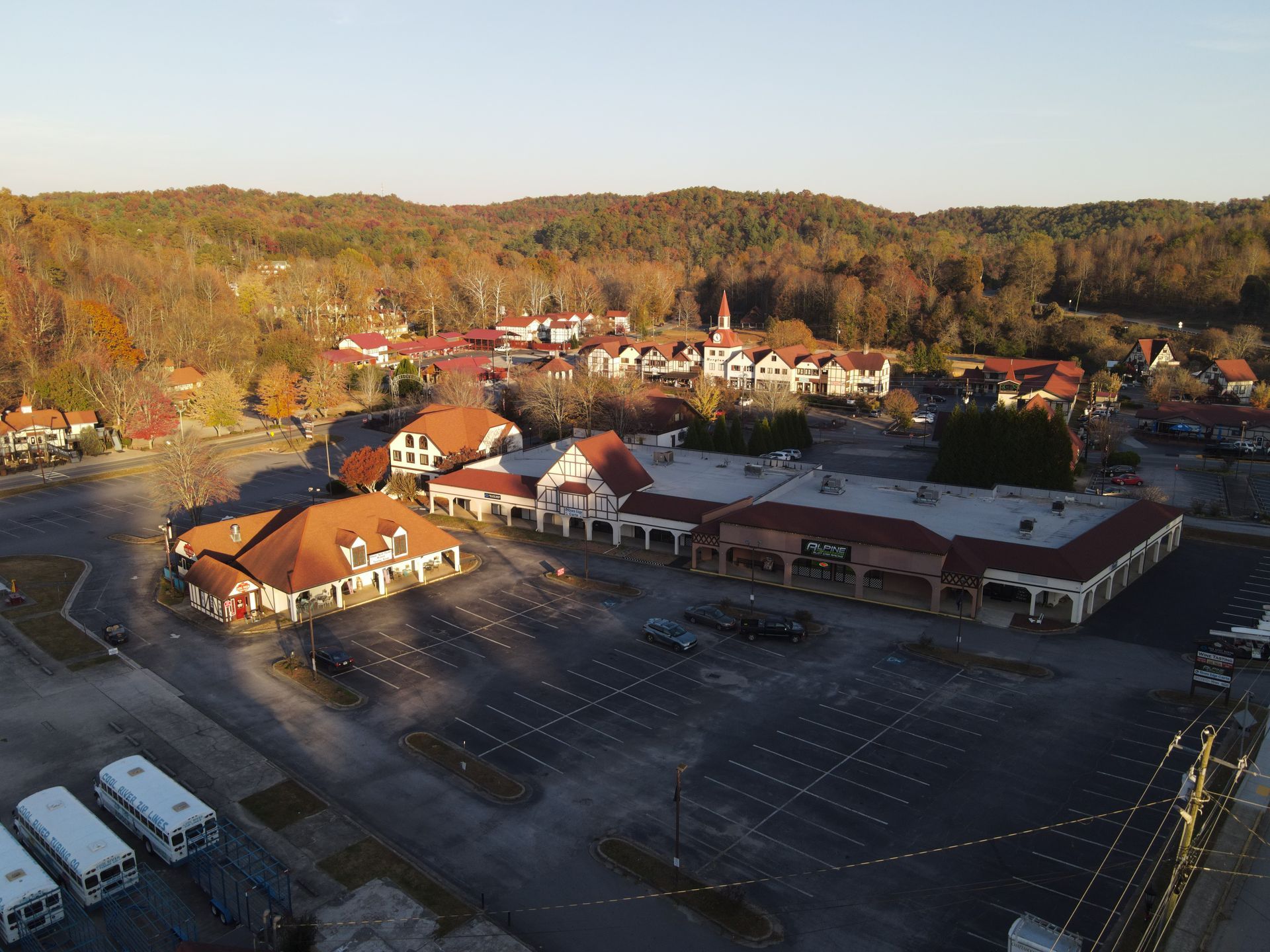 Aerial view of a commercial area with buildings, parking, and a forested hillside in autumn.