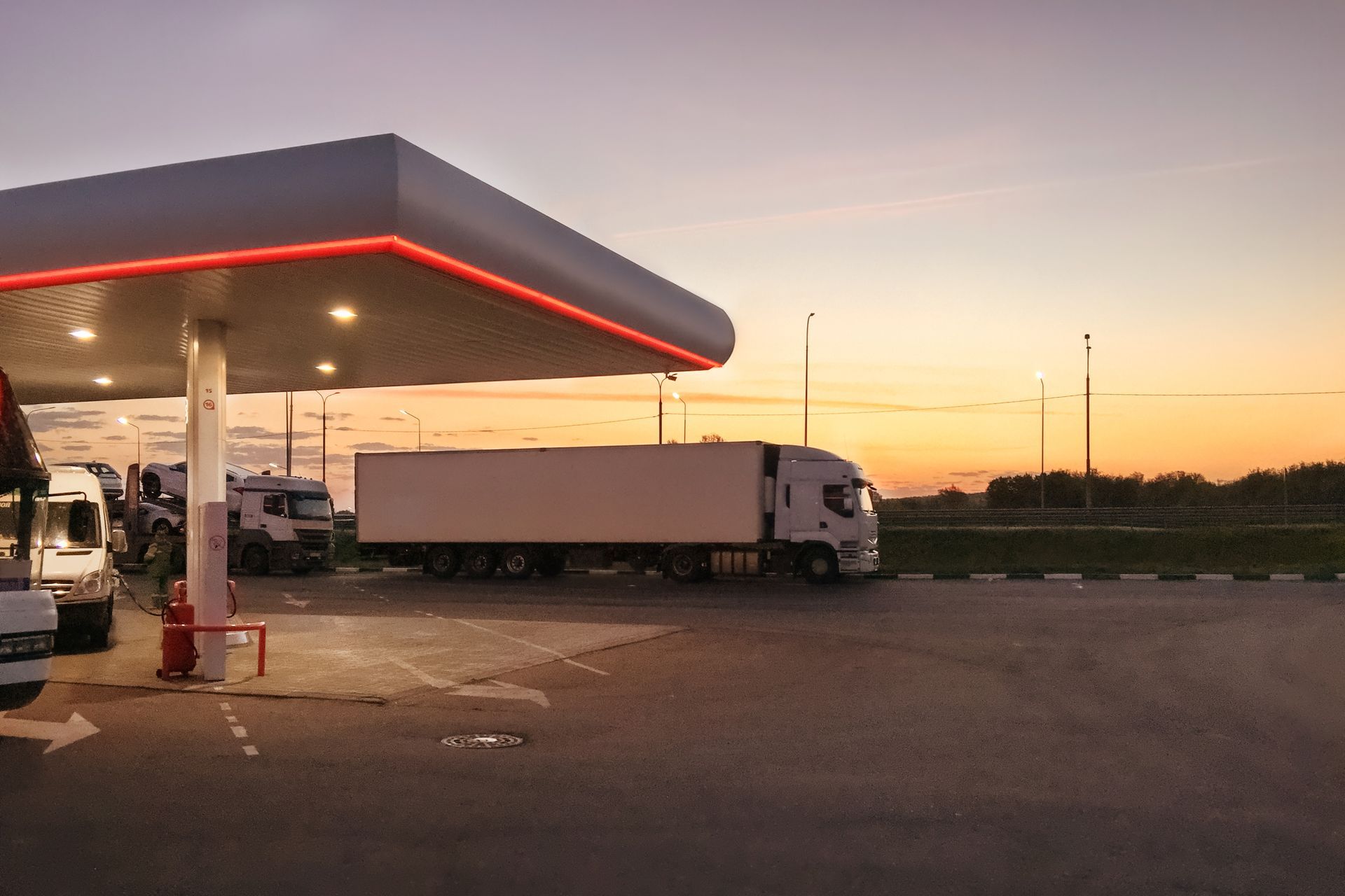 Trucks at a gas station under a canopy with red lights, at sunset.