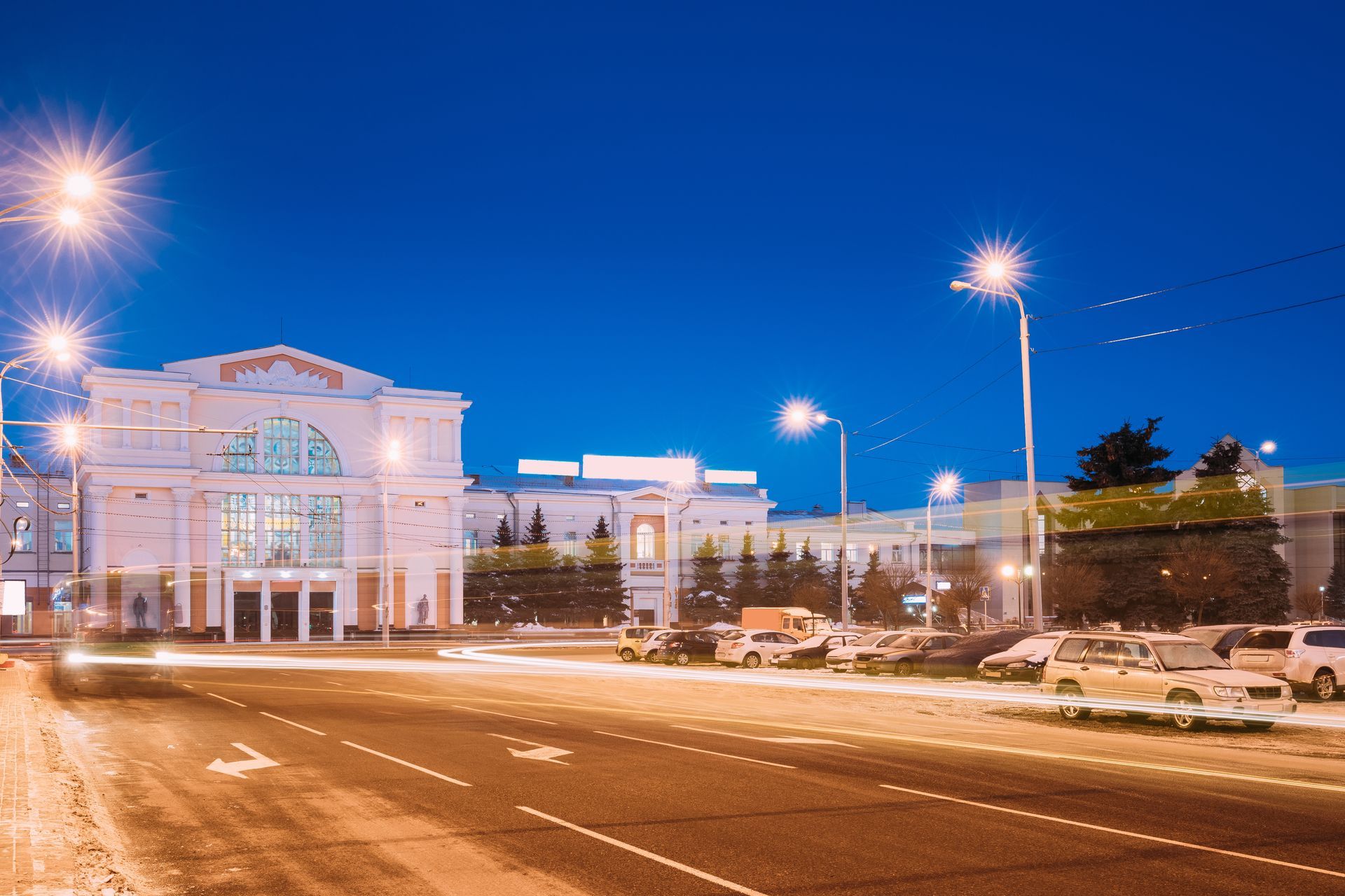 Night view of a grand white building with streetlights and passing cars on a dark blue sky background.