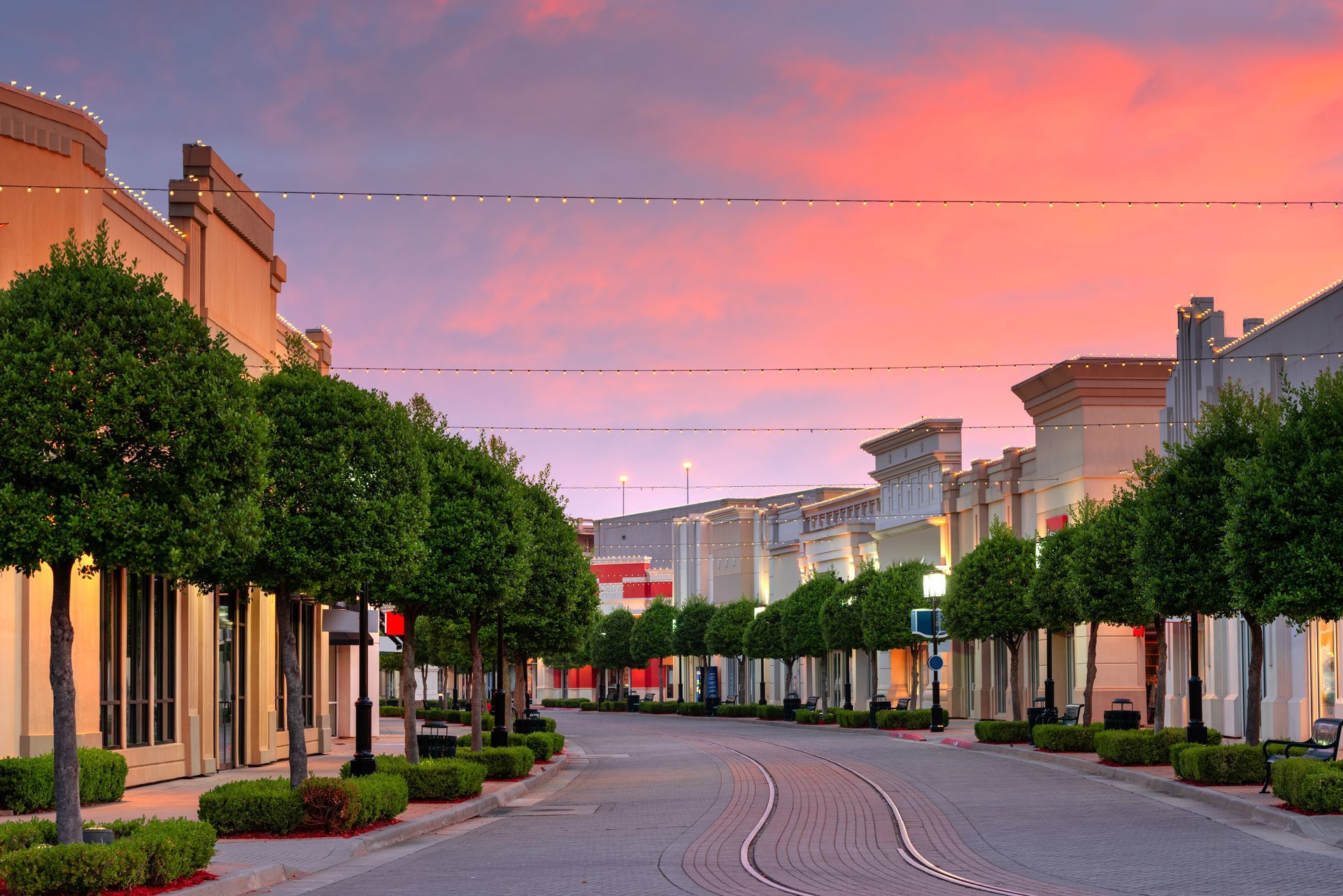 Cobblestone street lined with trees and shops at sunset; pink and orange sky.