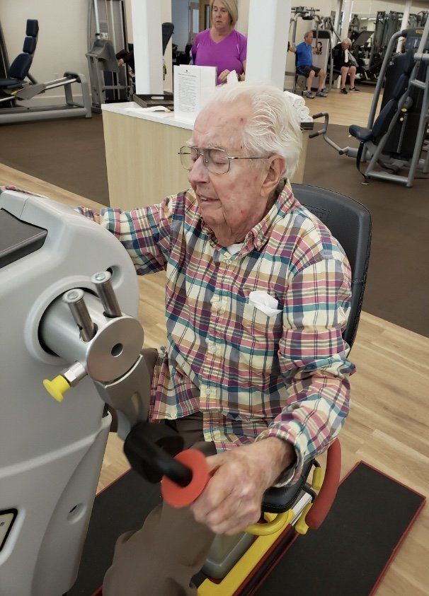 Older man using exercise machine in a gym. He is wearing glasses and a plaid shirt. A woman watches in the background.