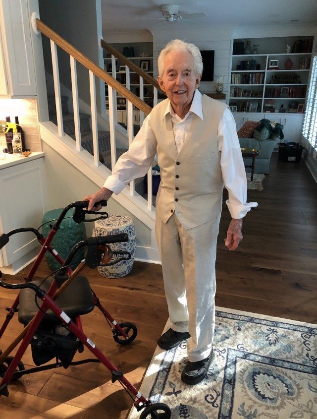 Elderly man in a tan suit standing next to a red walker inside a house with wooden floors.