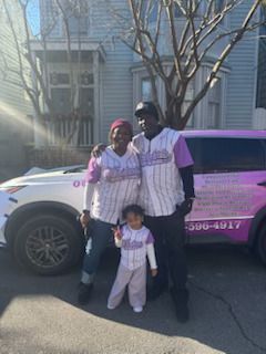 A family, in matching shirts, stands by a vehicle in front of a house on a sunny day.