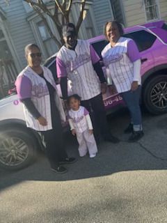 Group of four people, all wearing matching purple and white baseball jerseys, standing in front of two cars.