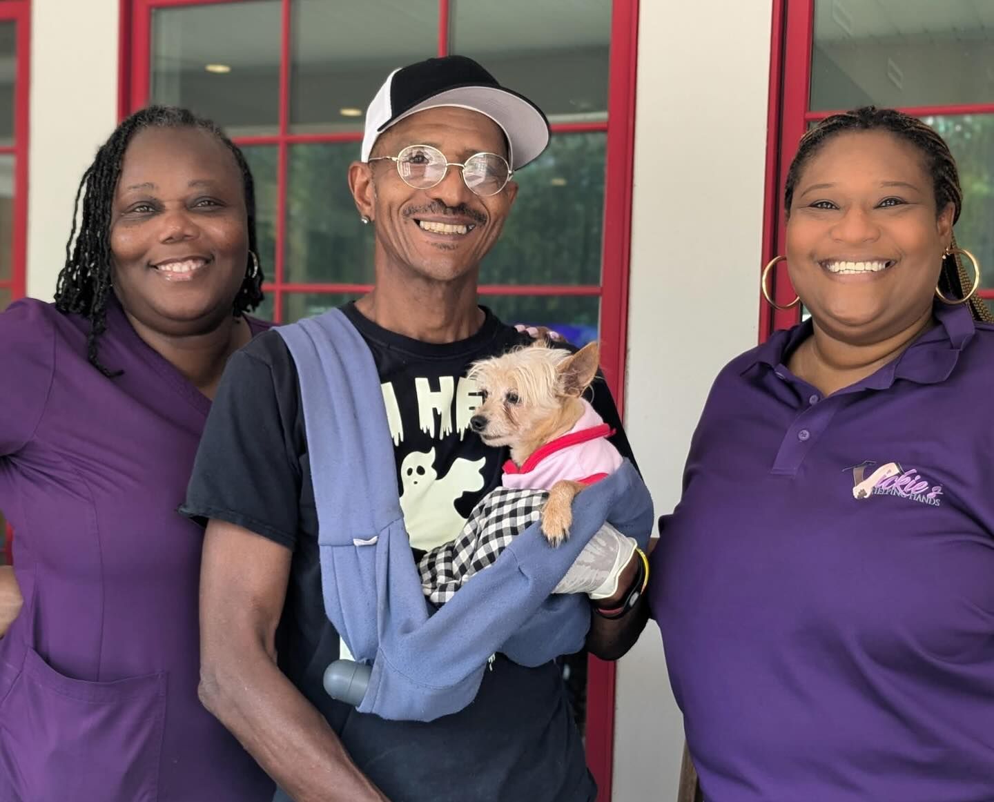Three people smiling, one holding a small dog in a blue carrier. Outdoor setting with a red-trimmed door.