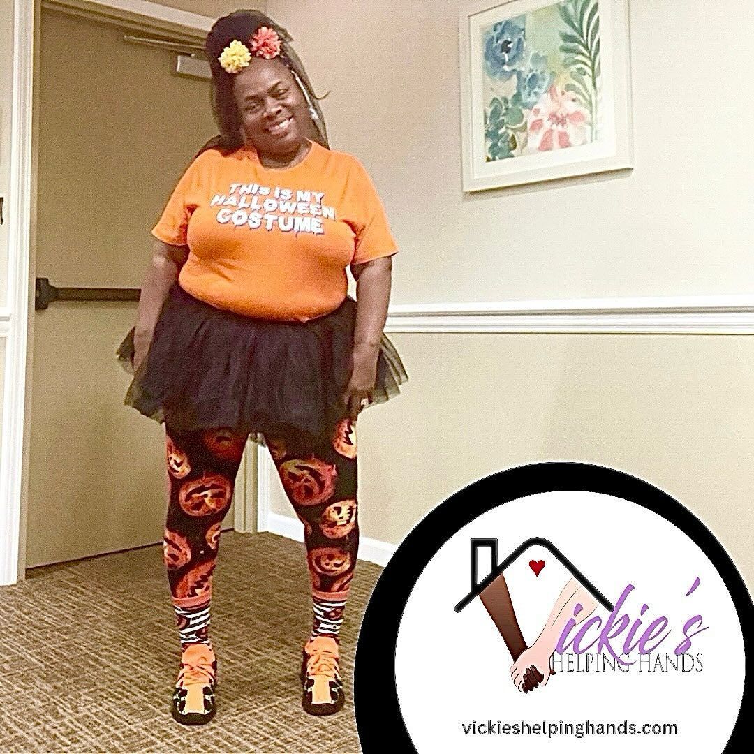 Woman in Halloween costume, orange shirt, black tutu, pumpkin leggings, orange shoes, smiling. Logo: Vickie's Helping Hands.