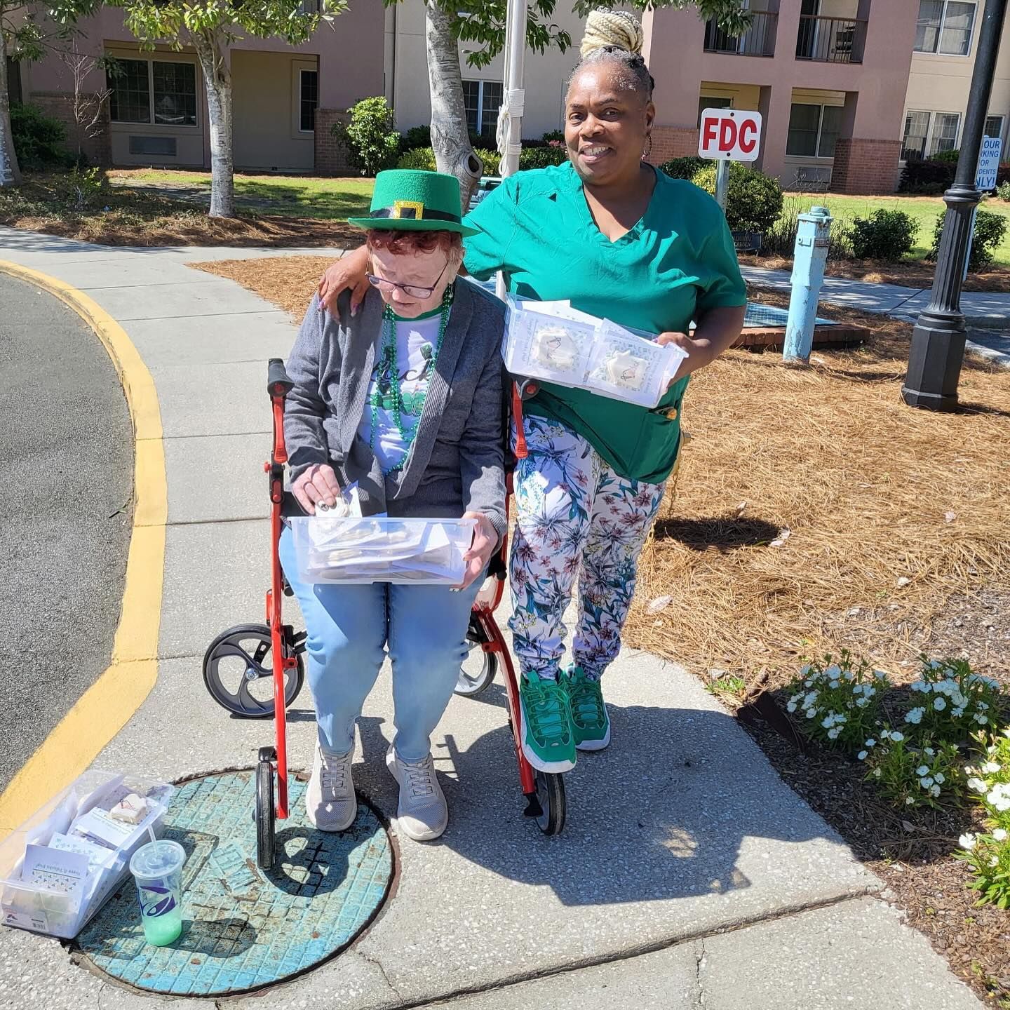 Two people on a sidewalk; one using a walker, both holding trays of food. One wears a green hat.