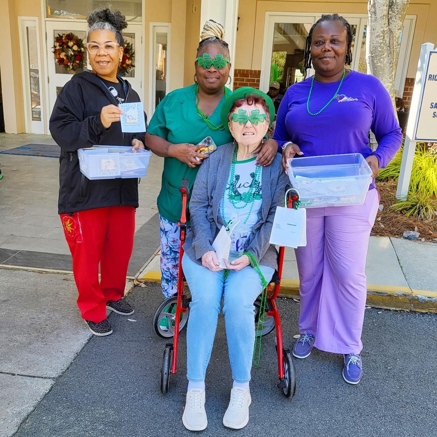 Four people in St. Patrick's Day attire holding baskets outside a building. One person is in a wheelchair.